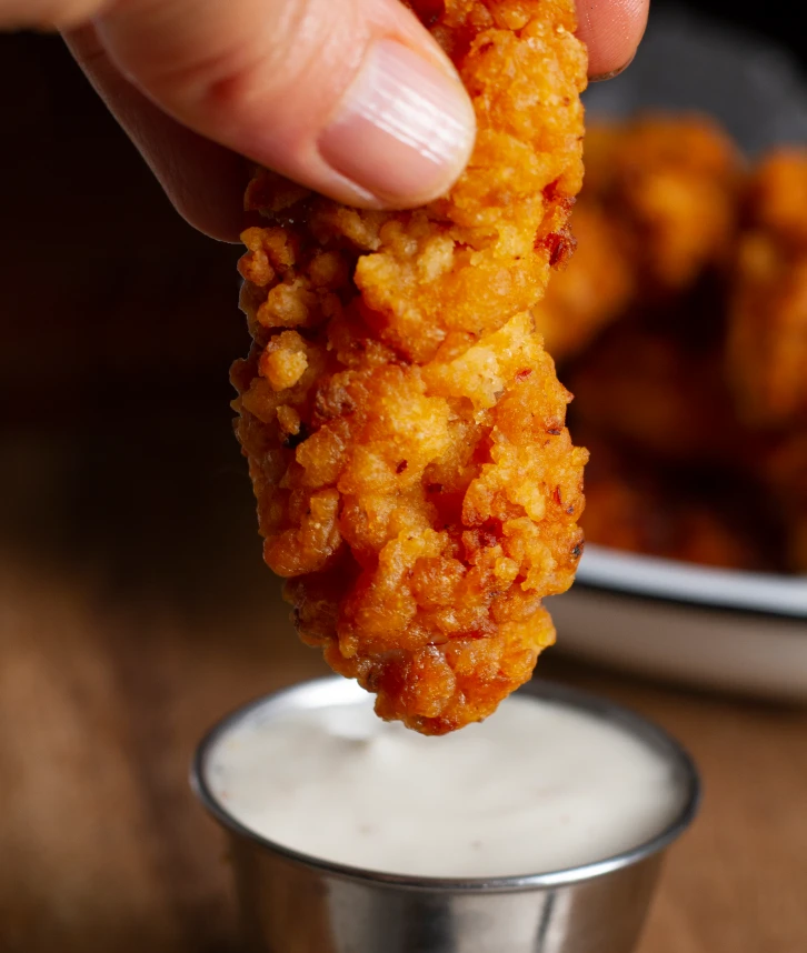 Un doigt de poulet croustillant tenu entre les doigts, prêt à être trempé dans une sauce blanche crémeuse dans un petit récipient en métal.