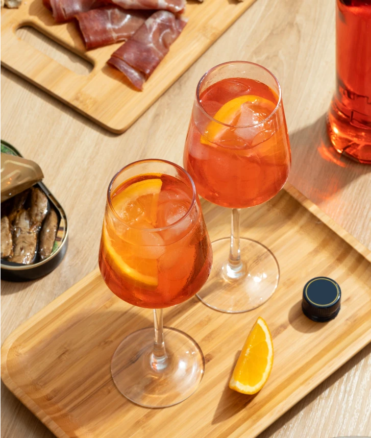 Deux verres de cocktail orange avec des glaçons et des tranches d’orange sont posés sur un plateau en bois. À l’arrière-plan, on voit une planche de charcuterie, une boîte de sardines ouverte et une bouteille de boisson orange.