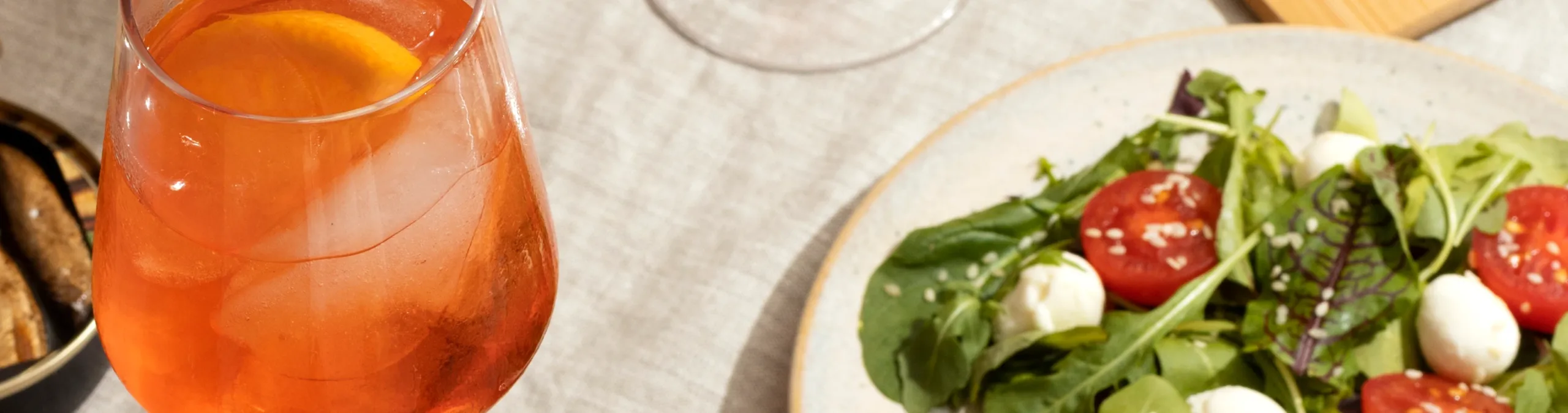 Gros plan d’un cocktail orange rafraîchissant avec des glaçons et une tranche d’orange, à côté d’une assiette de salade fraîche composée de tomates cerises, de billes de mozzarella et de verdure mélangée, le tout posé sur une nappe claire.