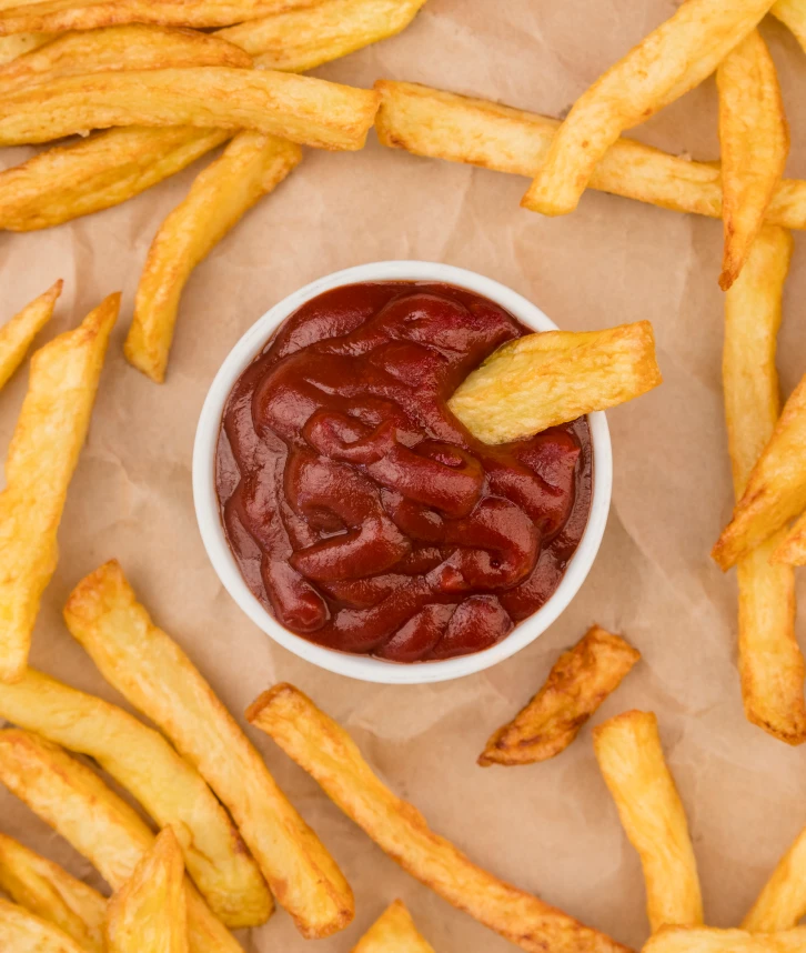 Des frites dorées disposées autour d’un petit bol de ketchup, avec une frite trempée dans la sauce, le tout sur un papier parchemin.