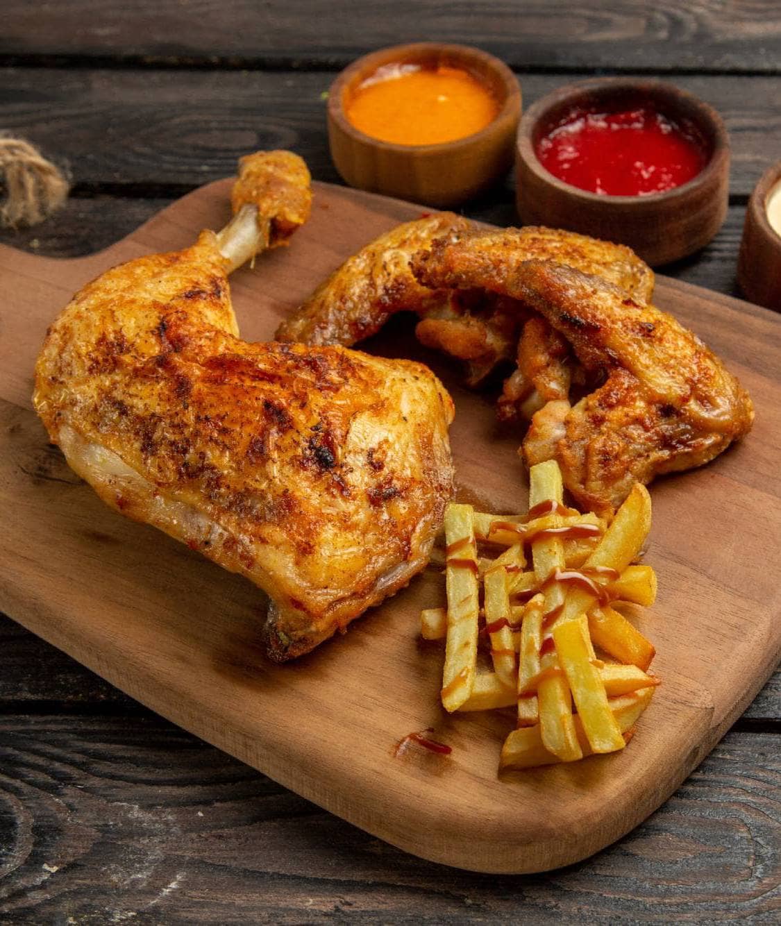 Vue rapprochée latérale de poulet, épices, bols, trois types de sauces, ailes et pilon de poulet avec frites, ketchup, sur une planche à découper, avec un fond sombre.